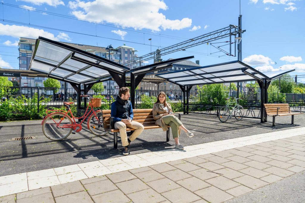 Dubbelsidigt cykeltak med parkbänk framför där en man och en kvinna sitter.
